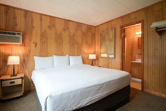 Wood-paneled hotel room with a king-size bed, white linens, and two bedside lamps; an air conditioner on the left wall and a doorway to an attached bathroom.