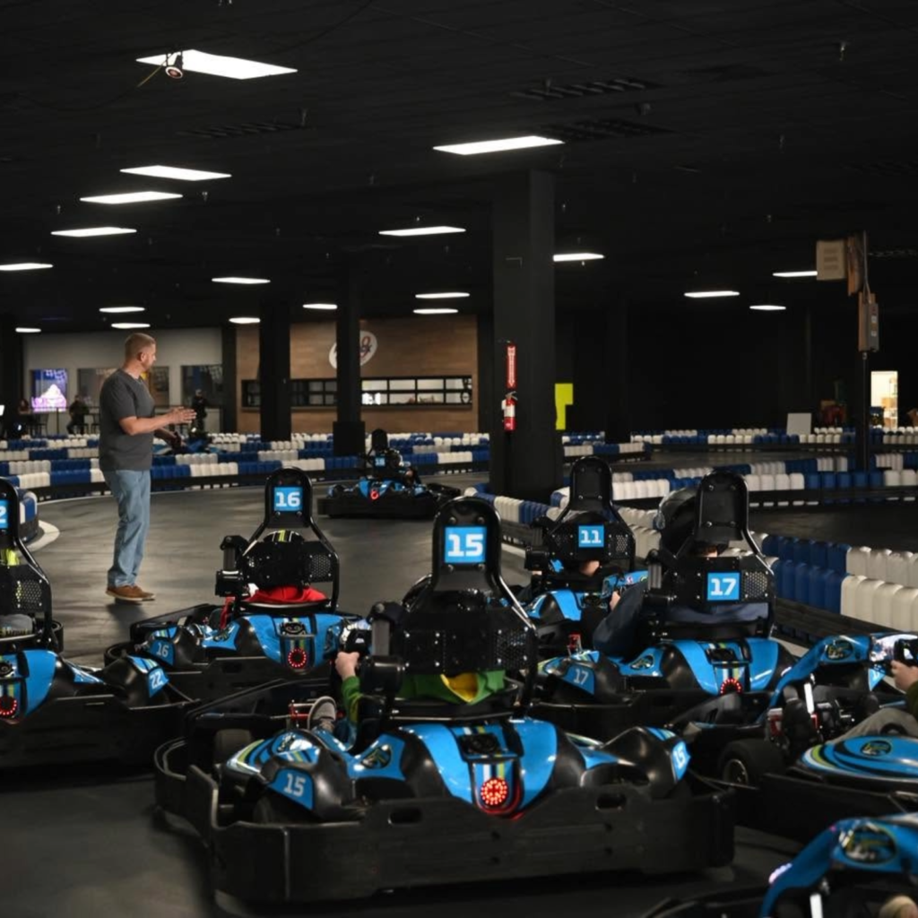 Indoor go-kart track with blue karts lined up and a man standing to the left, holding a helmet or controls.