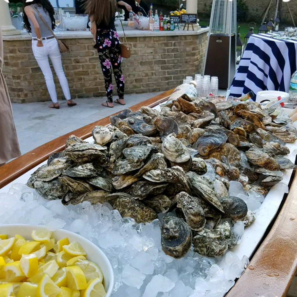 Pile of raw oysters on crushed ice at a seafood bar, with sliced lemons in a bowl nearby.