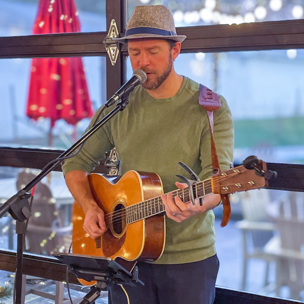 Male musician wearing a hat, playing an acoustic guitar and singing into a microphone outdoors.