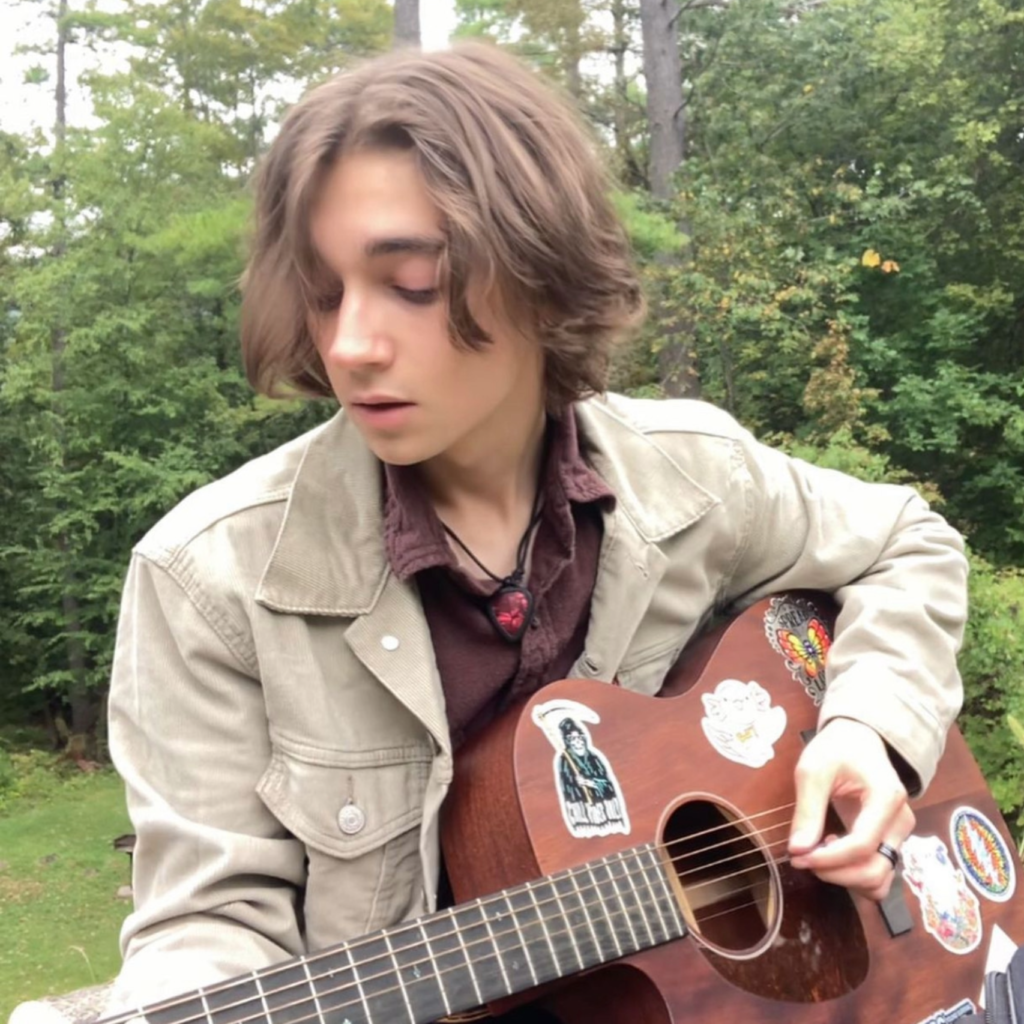 Young person sitting outdoors with a sticker-covered acoustic guitar, looking down at the strings.