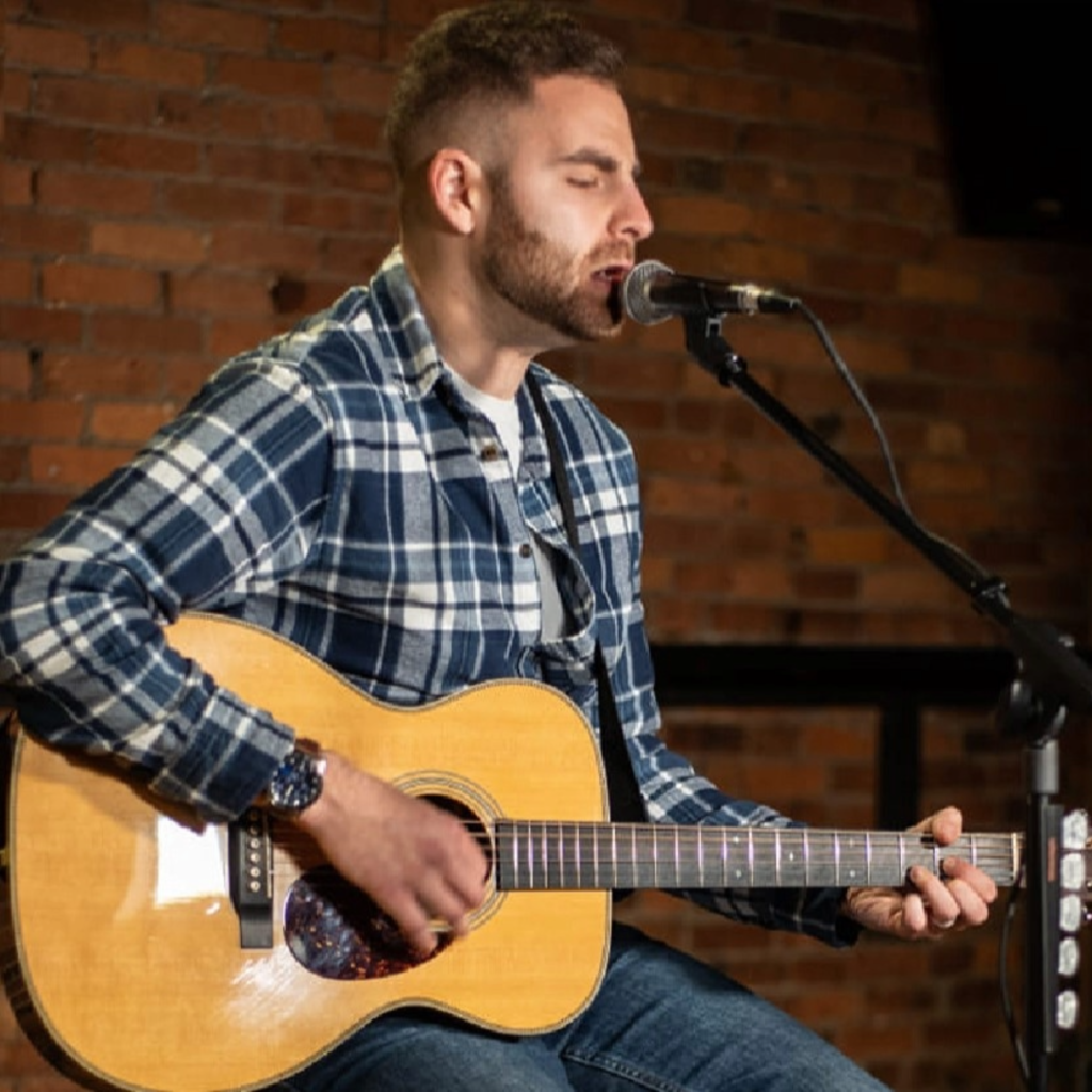 Male musician singing into a microphone while playing an acoustic guitar on stage, wearing a blue plaid shirt against a brick wall backdrop that suggests a small venue.