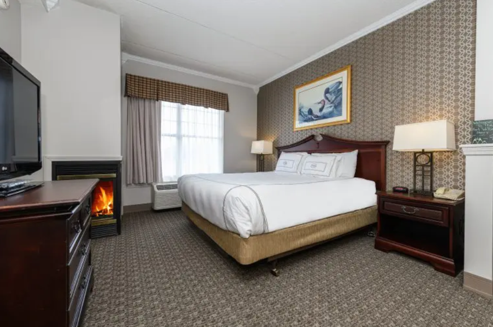 Hotel room with a king bed, two nightstands, lamps, a framed artwork, and a fireplace beside the window.