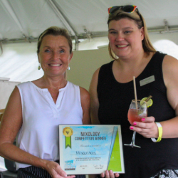 Two smiling women under a canopy, one holding a framed award and the other a cocktail, at a mixology competition winners event.