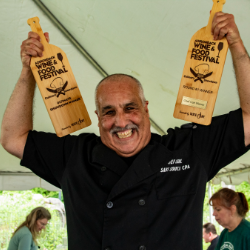 Smiling chef in a black uniform holding up two wooden bottle-shaped awards reading 'Wine & Food Festival'.
