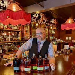 Bartender in a vest and tie behind a wooden bar with bottled liquor and red lamps in a warmly lit bar setting.