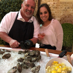 Man in an apron and a smiling woman hold wine glasses at a seafood bar, with a tray of oysters on ice and a bowl of lemon wedges in front.