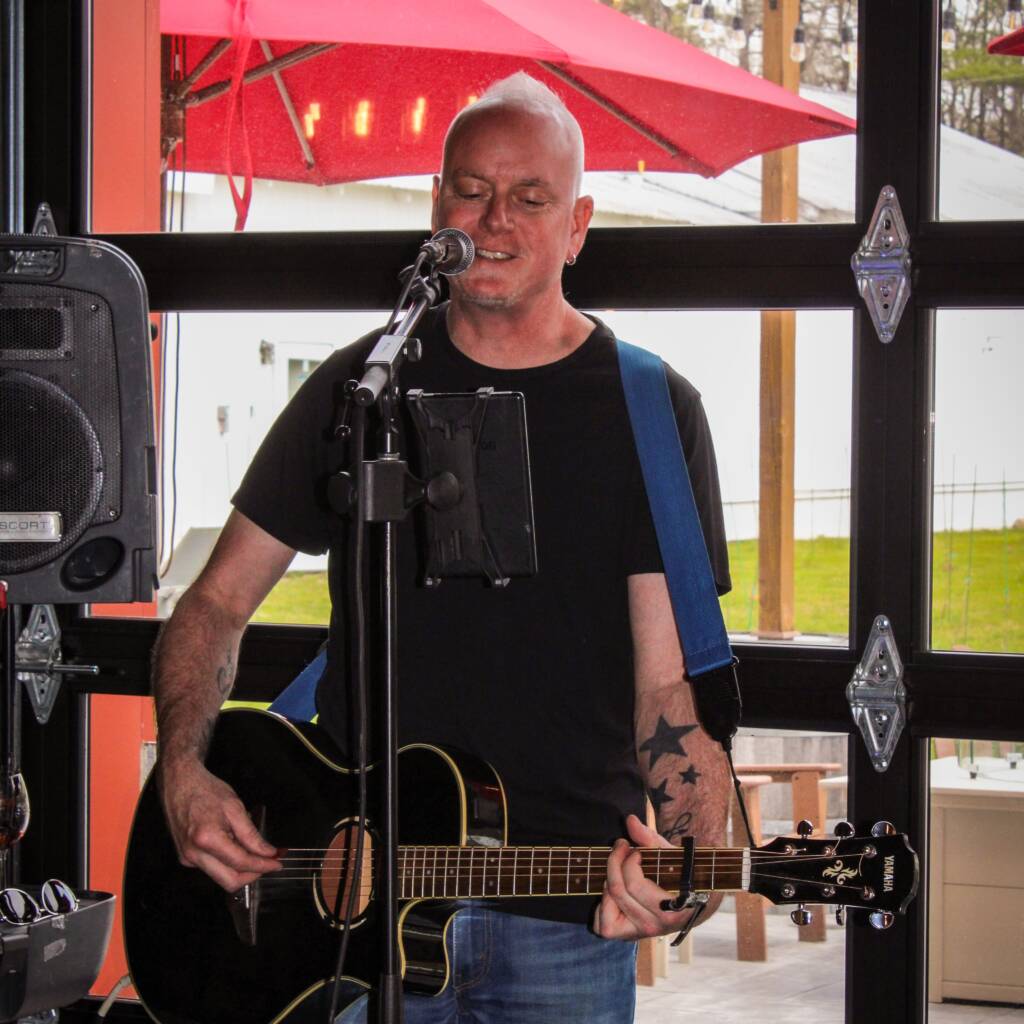 Male street musician playing an acoustic guitar and singing into a microphone, wearing a black shirt and blue suspenders, inside a glass booth with red exterior scenery behind him.