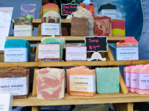 Assorted colorful handmade soaps on wooden shelves at a market, with a 'Soap $9' sign in the background.