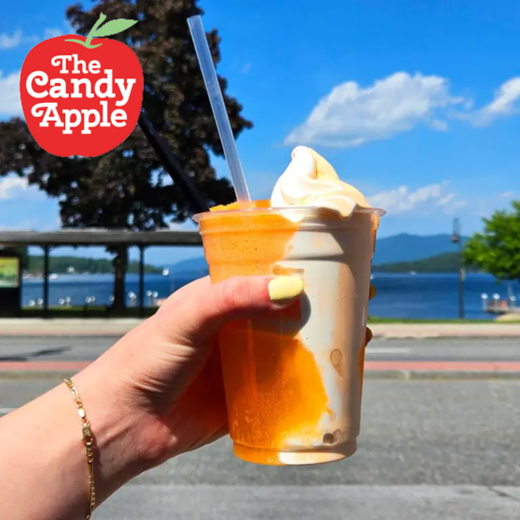 Hand holding a two-toned iced drink with whipped cream near a lake; The Candy Apple logo appears in the top-left corner.