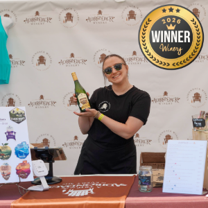 Woman in sunglasses at a winery booth holds up a bottle of sparkling wine, with a 2026 Winner Winery badge in the backdrop.