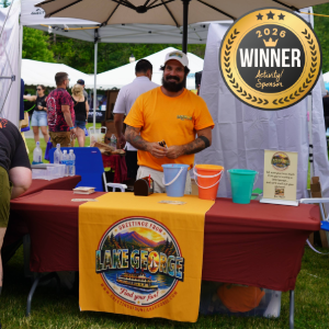 Man in an orange shirt behind a Lake George festival booth, with colorful buckets on the table and a 2026 winner badge visible in the foreground.
