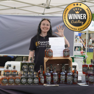 Smiling woman at an outdoor booth waves and holds a certificate behind a display of bottled spirits and a 2023 Winner badge on a tent.