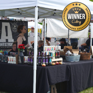 Outdoor market stall selling bottles of cider under a canopy, with a large “Winner 2026 Cider” badge visible above the table.