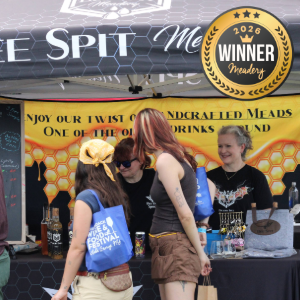 People at a festival beverage booth; staff in blue vests serve drinks behind a counter, while a woman in brown shorts talks with them, with a yellow honeycomb banner in the background.