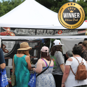 People line up at an outdoor tent booth with a 'Cheese Filled Company' banner and a large 'WINNER 2026' artisan food badge above the stall.