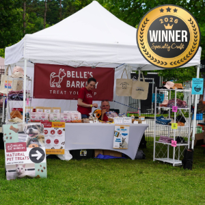 Outdoor market stall under a white canopy selling pet treats; banner reads 'Belle's Bark'; vendor behind the table with products and bags, plus a gold winner badge in the top-right corner.