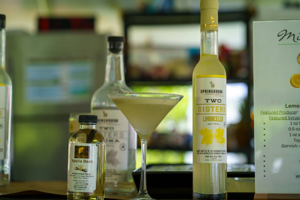 Bar shelf with a tall lemon liqueur bottle labeled Springbrook and a cocktail glass in front of it, blurred bottles in the background.