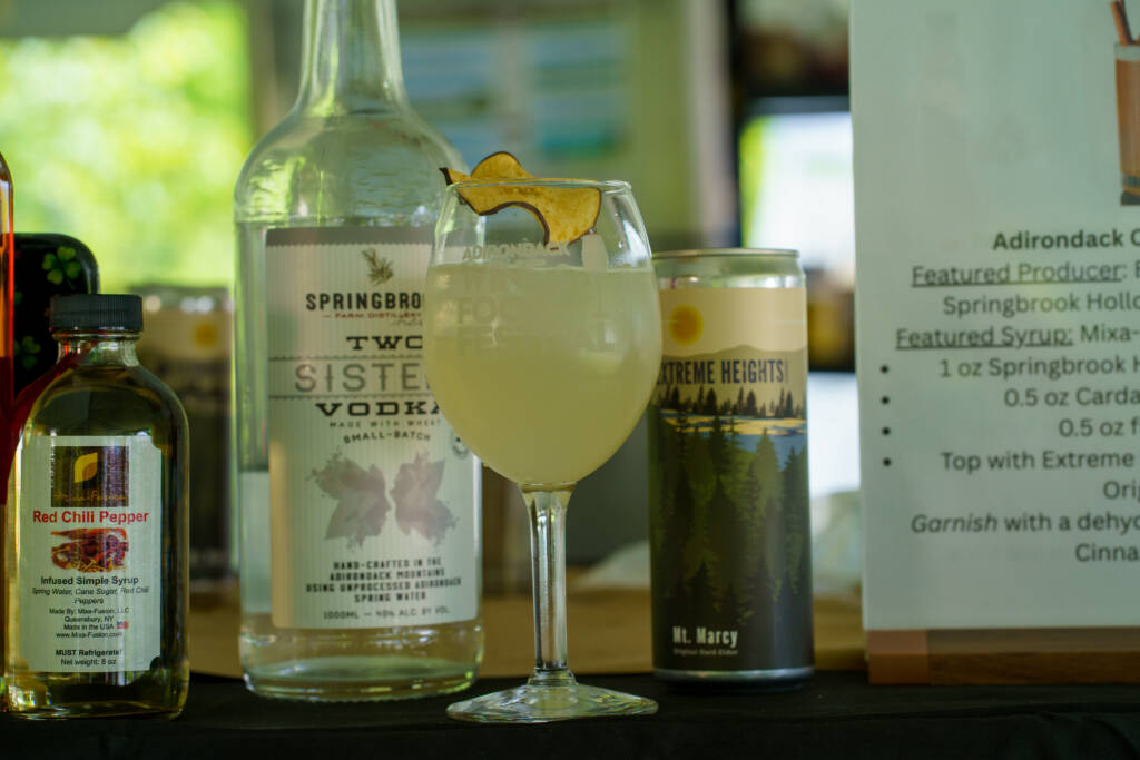 Stemmed cocktail glass with a pale drink and a lemon twist, surrounded by liquor bottles and a menu board in the background.