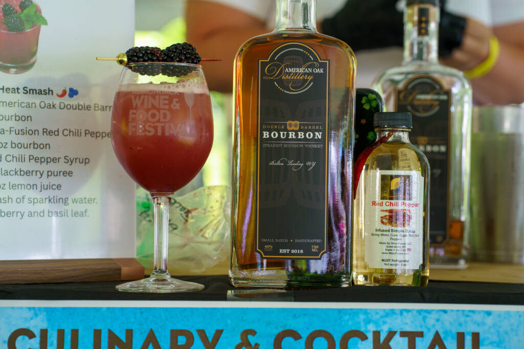 Bourbon bottle from American Oak Distillery beside a red cocktail garnished with blackberries on a skewer at a bar.