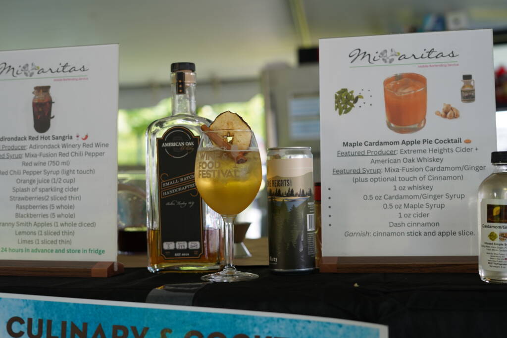 Cocktail with orange juice and a pear-like garnish in a stemmed glass, flanked by festival posters and a whiskey bottle nearby.