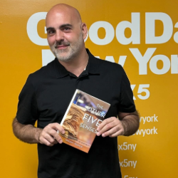 Man in a black shirt holding a book titled 'Five Senses' in front of a bright yellow event backdrop with branding text.