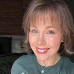 Portrait of a smiling woman with short blonde-brown hair wearing a green shirt, indoor bedroom backdrop with pillows behind her.